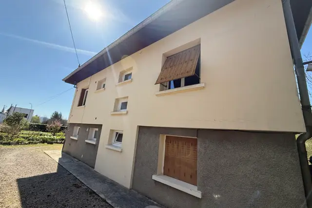 Façade d'une maison crépie en jaune pâle, fissures dans l'enduit, volets en bois, soubassement en crépi gris, sous le soleil