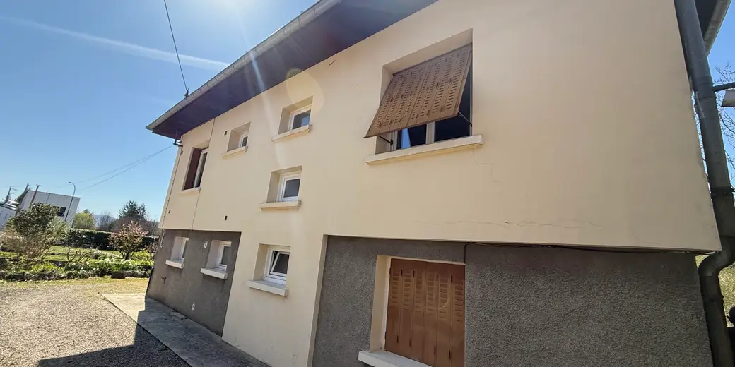 Façade d'une maison crépie en jaune pâle, fissures dans l'enduit, volets en bois, soubassement en crépi gris, sous le soleil