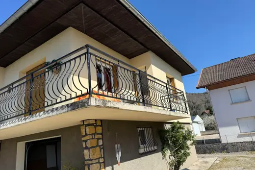 Vue d'ensemble de la maison avant travaux : balcon en fer forgé, avant-toit en bois, murs crépis clairs, soubassement en pierre et crépi gris, garage ouvert
