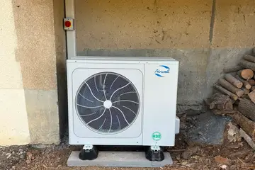 Unité extérieure blanche à un ventilateur, posée sur plots antivibratiles sur une dalle béton, sous un balcon, goulotte et interrupteur de coupure au mur