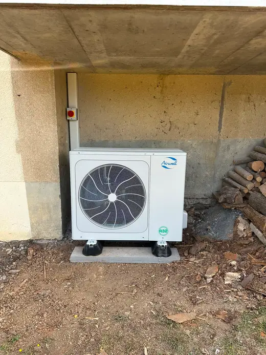 Unité extérieure blanche à un ventilateur, posée sur plots antivibratiles sur une dalle béton, sous un balcon, goulotte et interrupteur de coupure au mur