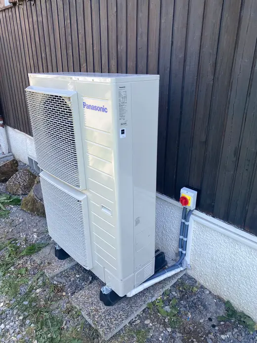 Unité extérieure beige à deux ventilateurs, contre un bardage en bois sombre, interrupteur de coupure rouge et jaune au mur, câbles sous gaine