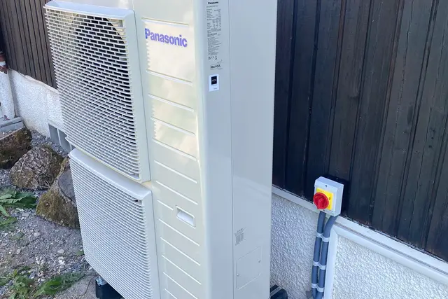 Unité extérieure beige à deux ventilateurs, contre un bardage en bois sombre, interrupteur de coupure rouge et jaune au mur, câbles sous gaine