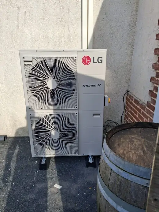 Unité extérieure à deux ventilateurs superposés, posée sur plots contre un mur crépi clair, mur de briques rouges et tonneau en bois à côté