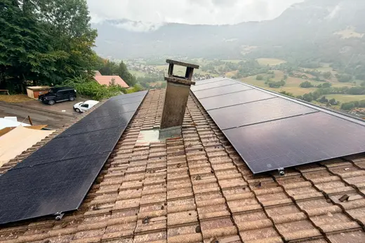 Deux groupes de panneaux photovoltaïques de part et d'autre d'une cheminée en béton, toiture en tuiles brunes, vallée et montagnes sous les nuages