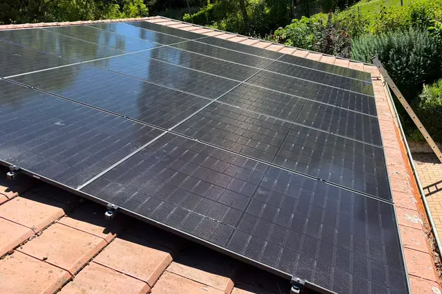 Trois rangées de panneaux photovoltaïques noirs sur une toiture en tuiles, jardin et haies en contrebas