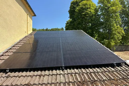 Dix panneaux photovoltaïques noirs en deux rangées sur une toiture en tuiles brunes, mur crépi jaune et grands arbres