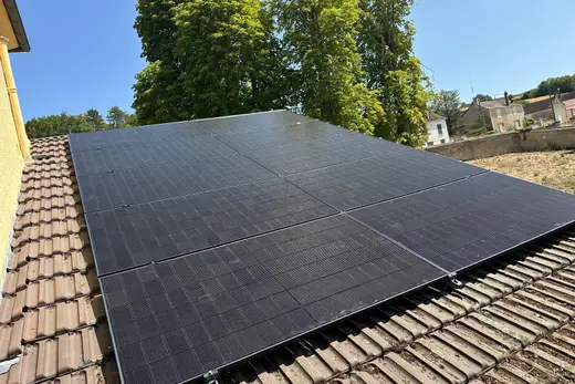 Dix panneaux photovoltaïques vus en oblique sur une toiture en tuiles brunes, maisons de pierre et arbres à l'arrière-plan