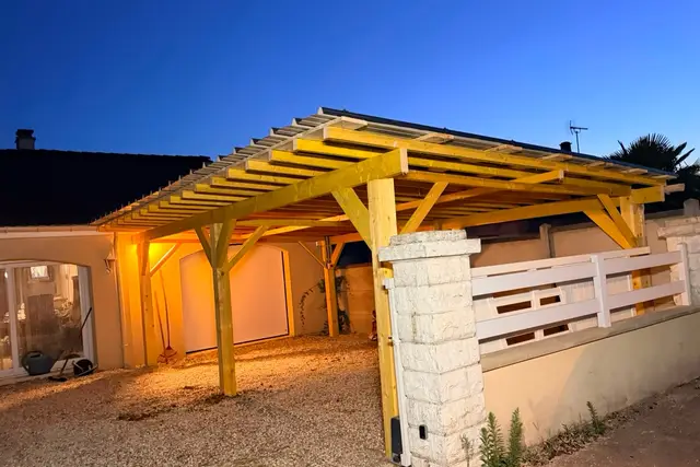 Carport en bois à deux places devant la maison et son garage, poteaux et contrefiches, toiture éclairée à la tombée du jour
