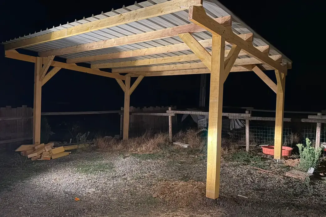 Abri en bois à quatre poteaux avec contrefiches, toiture en bac acier, photographié de nuit à la lampe, clôture en bois derrière
