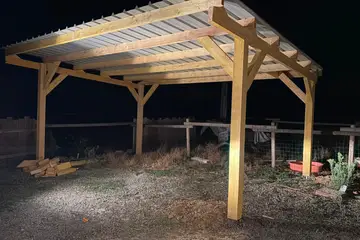 Abri en bois à quatre poteaux avec contrefiches, toiture en bac acier, photographié de nuit à la lampe, clôture en bois derrière