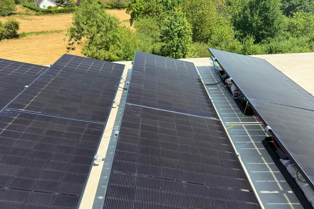 Gros plan des trois rangées de panneaux inclinés sur le toit plat, câblage et micro-onduleurs visibles sous le dernier rang, campagne en fond