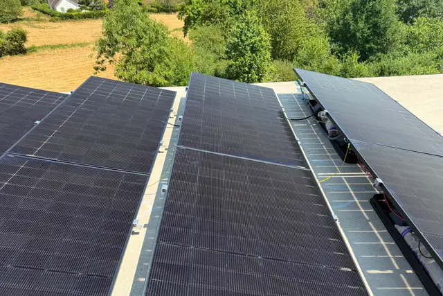 Gros plan des trois rangées de panneaux inclinés sur le toit plat, câblage et micro-onduleurs visibles sous le dernier rang, campagne en fond