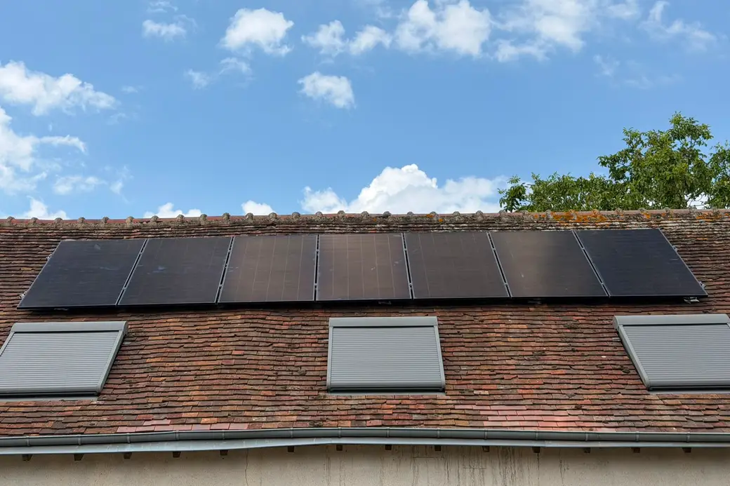 Sept panneaux photovoltaïques près du faîtage d'une toiture en tuiles plates anciennes, au-dessus de trois fenêtres de toit à volet
