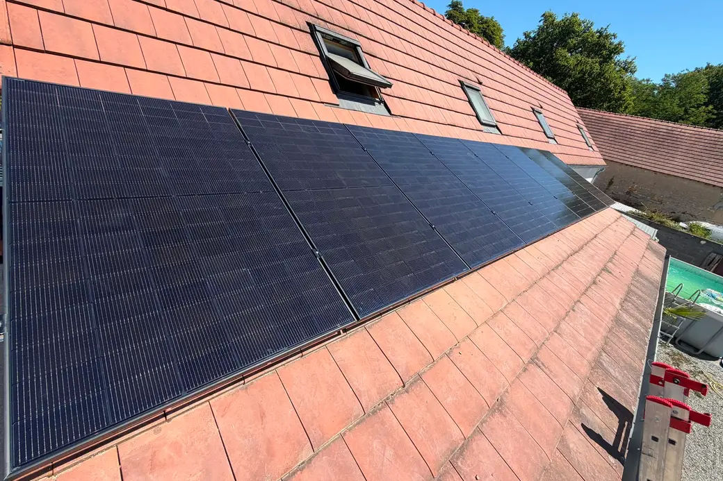Rangée de huit panneaux photovoltaïques au bas d'une toiture en tuiles rouges, trois fenêtres de toit, forêt en arrière-plan