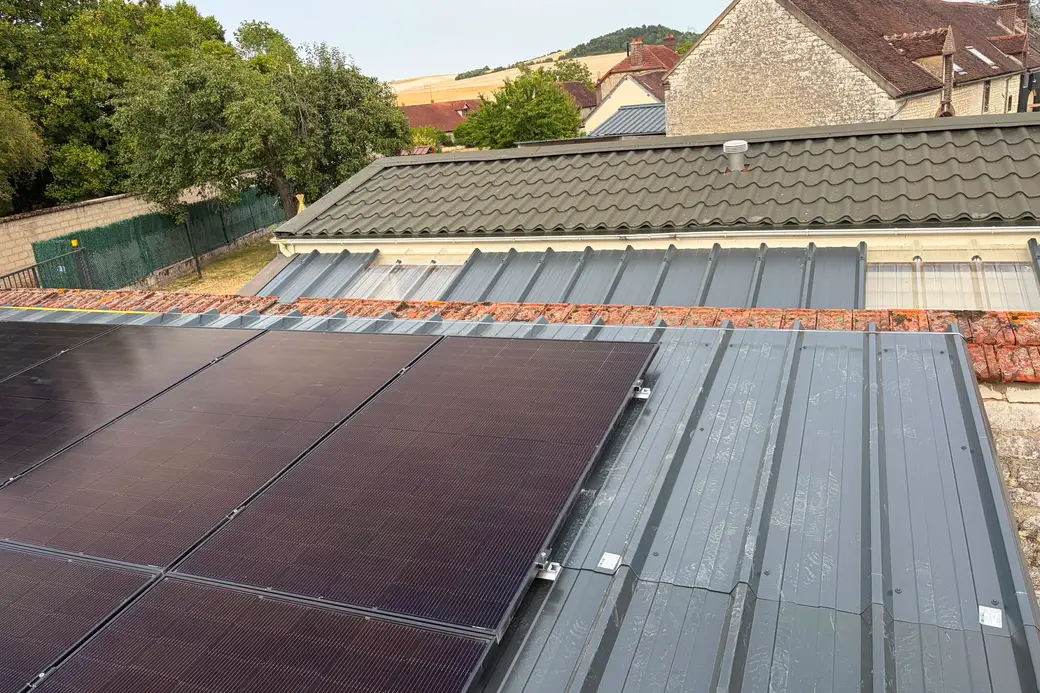 Panneaux photovoltaïques sur une toiture en bac acier gris, faîtage en vieilles tuiles, village de maisons en pierre et colline